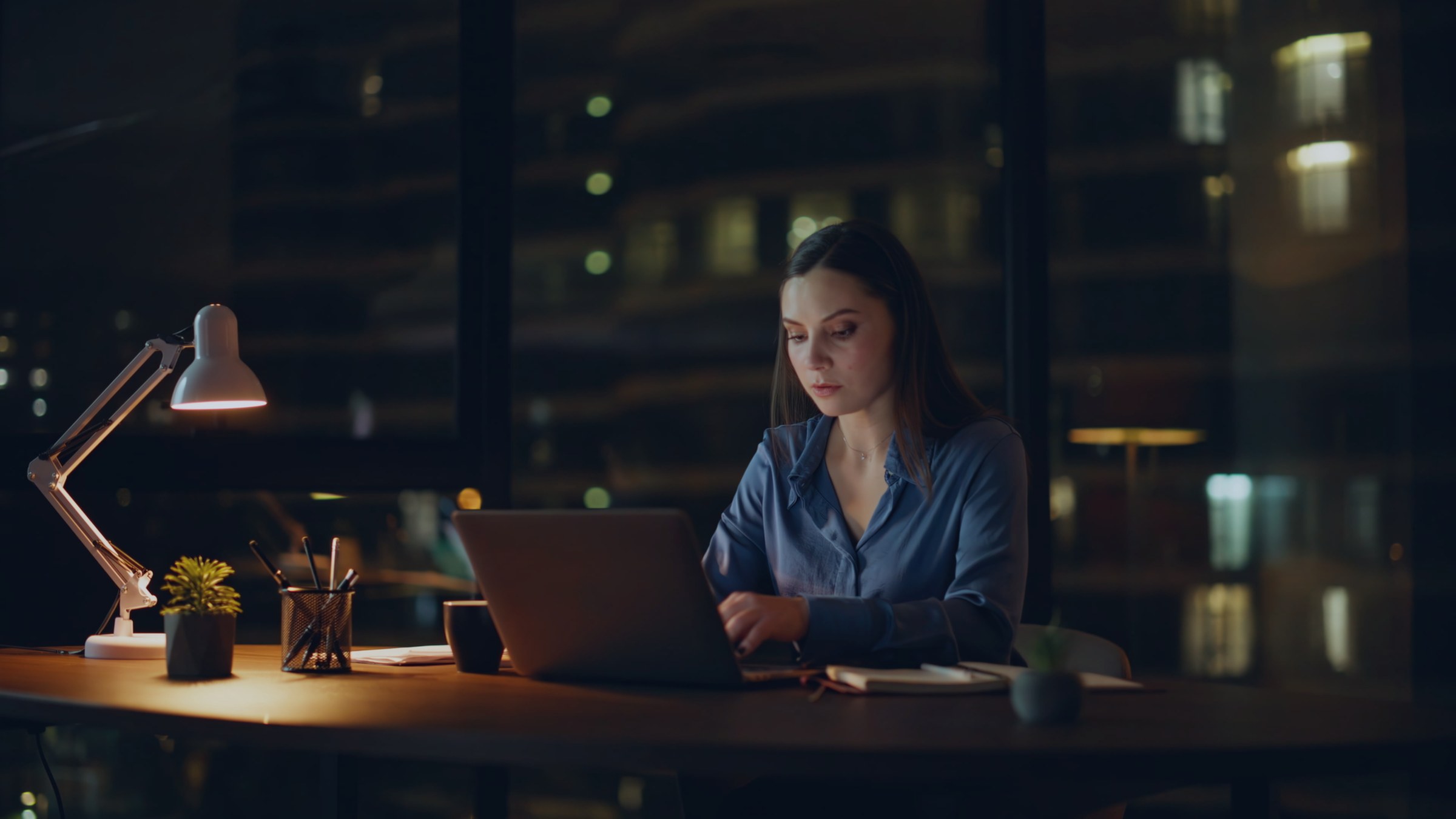 A woman working at a laptop late in the evening, city lights behind her.
